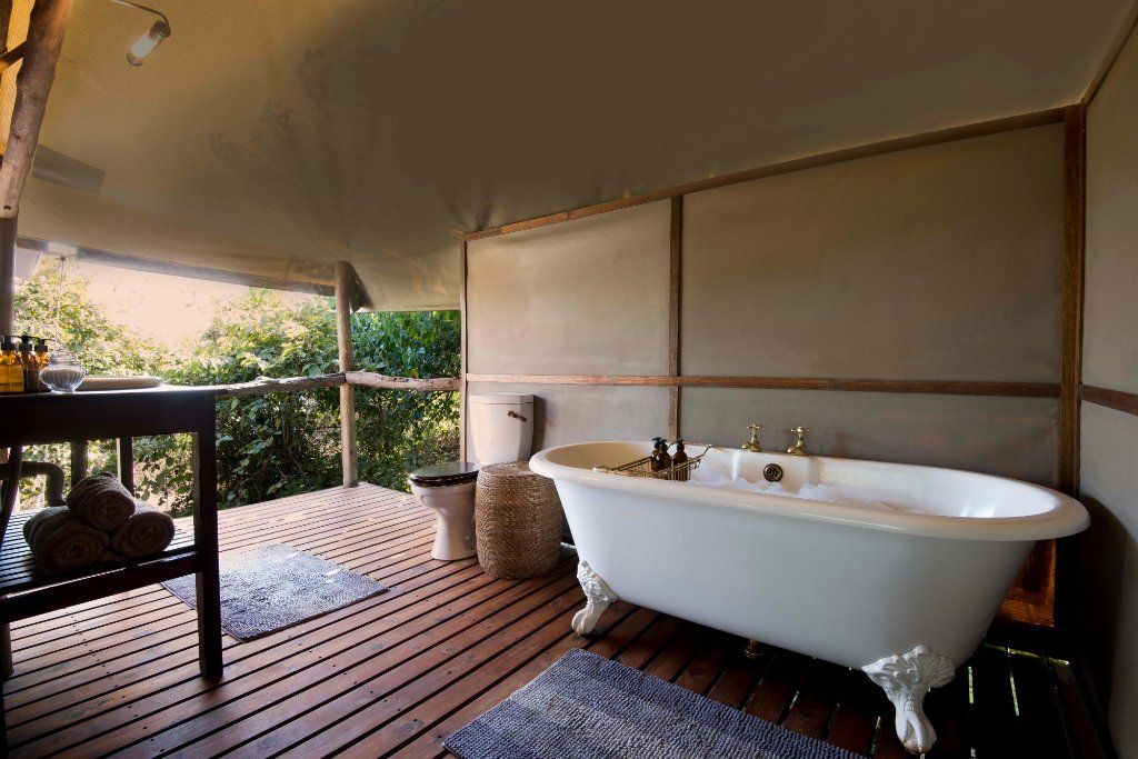 Outdoor bathroom with a claw-foot tub, toilet, wooden deck, and open view of trees.