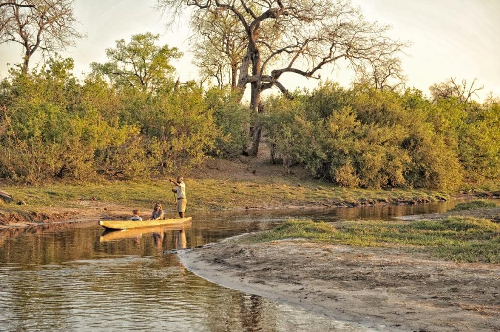 A small boat with three people on a river, surrounded by green trees and bushes, under a light sky.