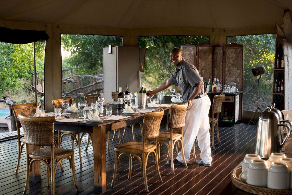 Waiter setting a dining table in an open-air tent with view of trees and pool.
