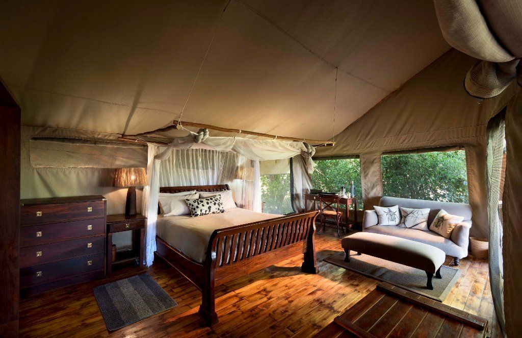 Luxury safari tent interior with wooden furniture, bed, and sofa; windows reveal lush greenery.