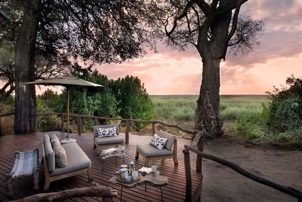Wooden deck with seating overlooking a savanna at sunset, trees frame the view.
