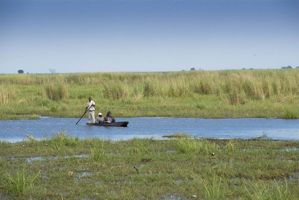 A small boat with three people on a calm waterway, surrounded by marshland under a blue sky.