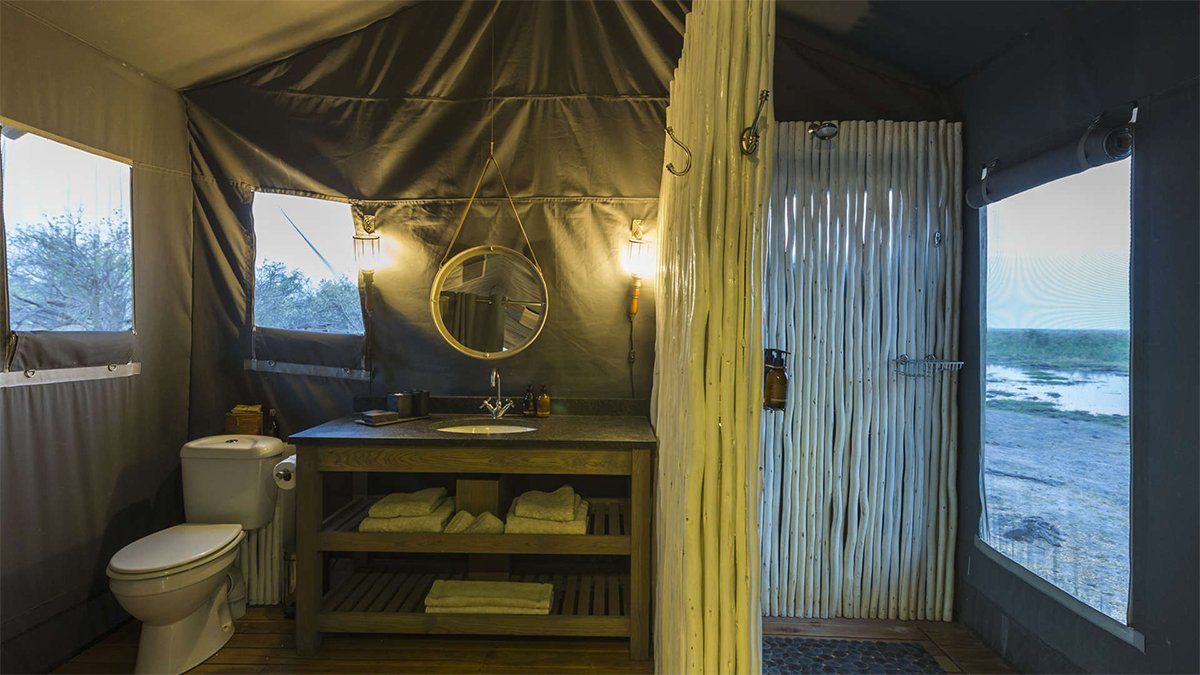 Bathroom in safari tent with toilet, vanity, and shower. Windows offer a view.