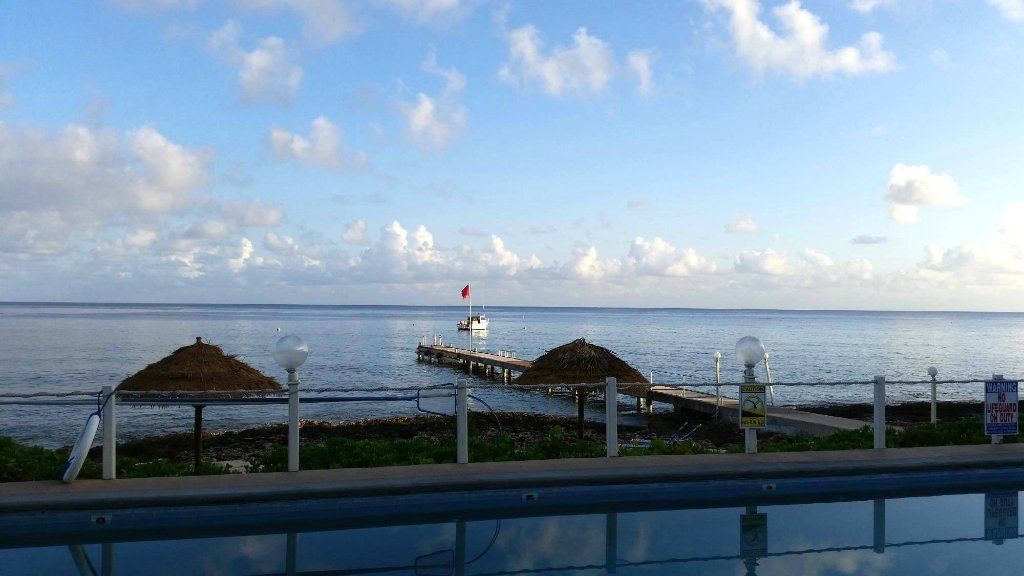 Ocean view with pier, thatched umbrellas, and swimming pool reflection under a cloudy sky.