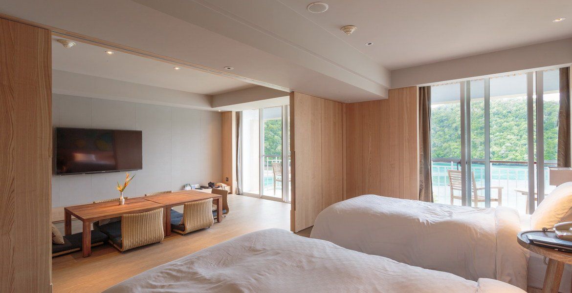 Modern hotel room with two beds, a table, and a view of a pool. Light wood and white bedding.