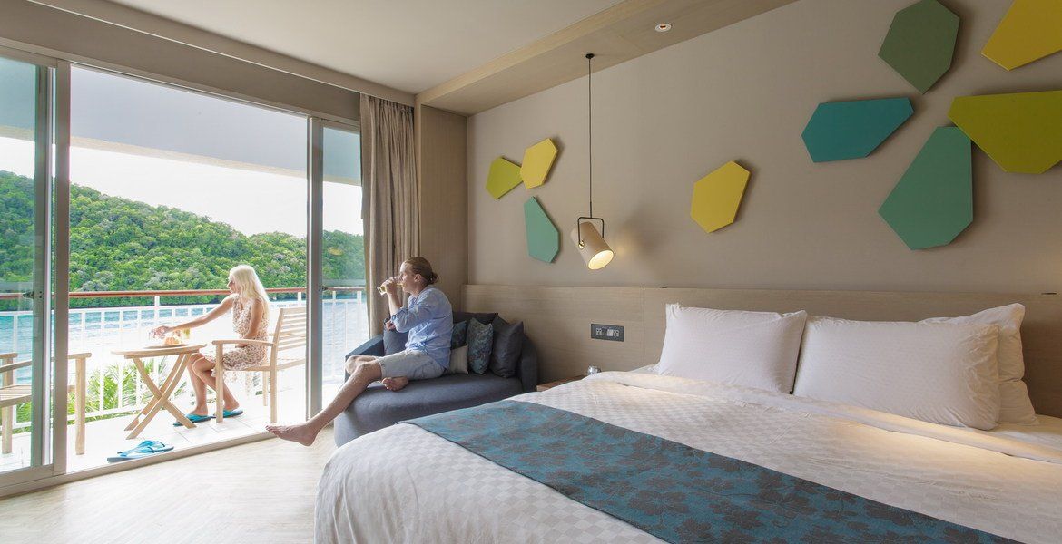 Hotel room with bed, balcony overlooking water, people relaxing.