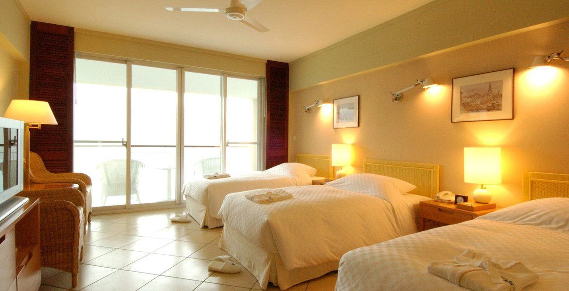 Hotel room with three beds, TV, and balcony access. Beige and light brown tones, with bright lighting.