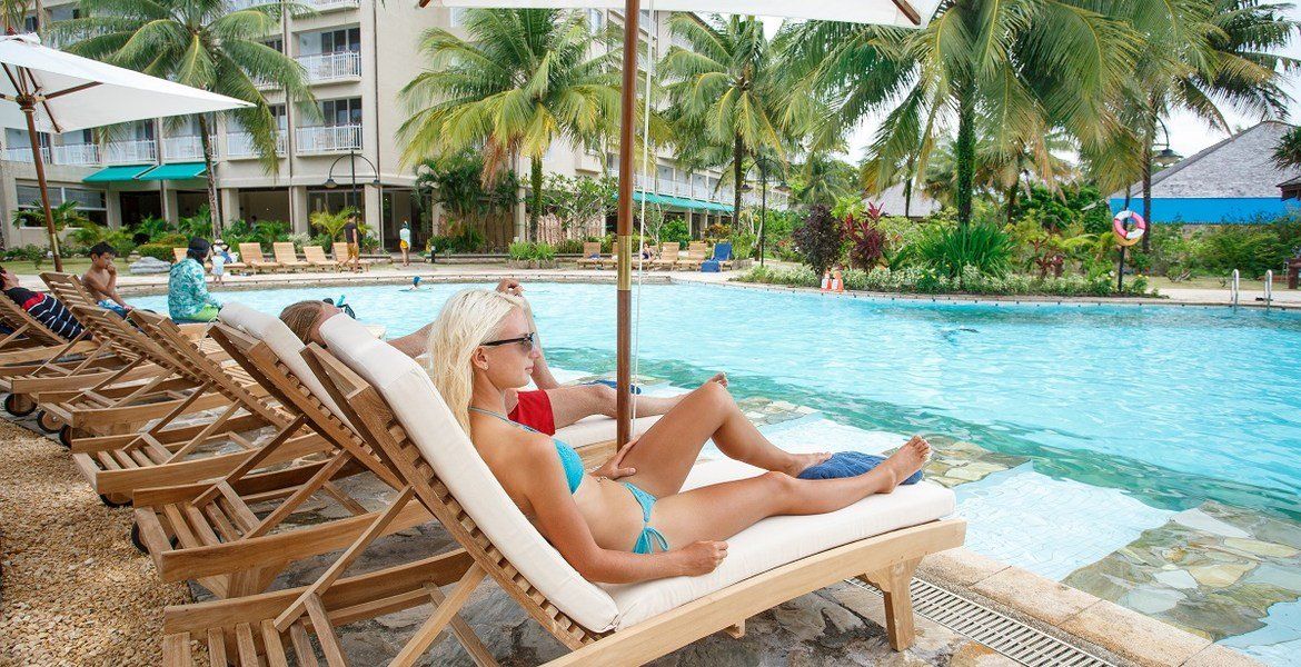 Woman in blue bikini relaxes on a poolside lounge chair under an umbrella.