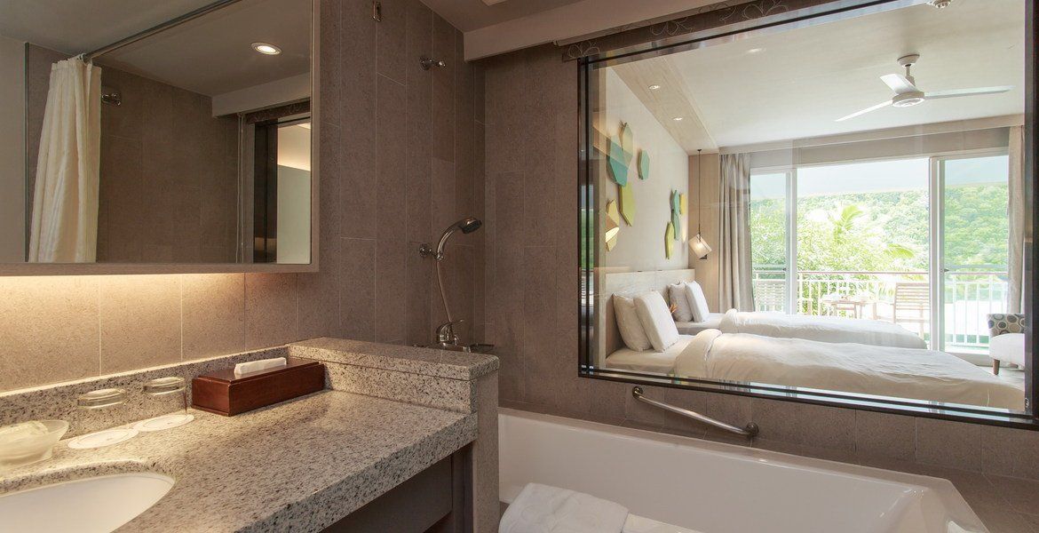 Bathroom with large mirror reflecting a bedroom with two beds and a balcony.
