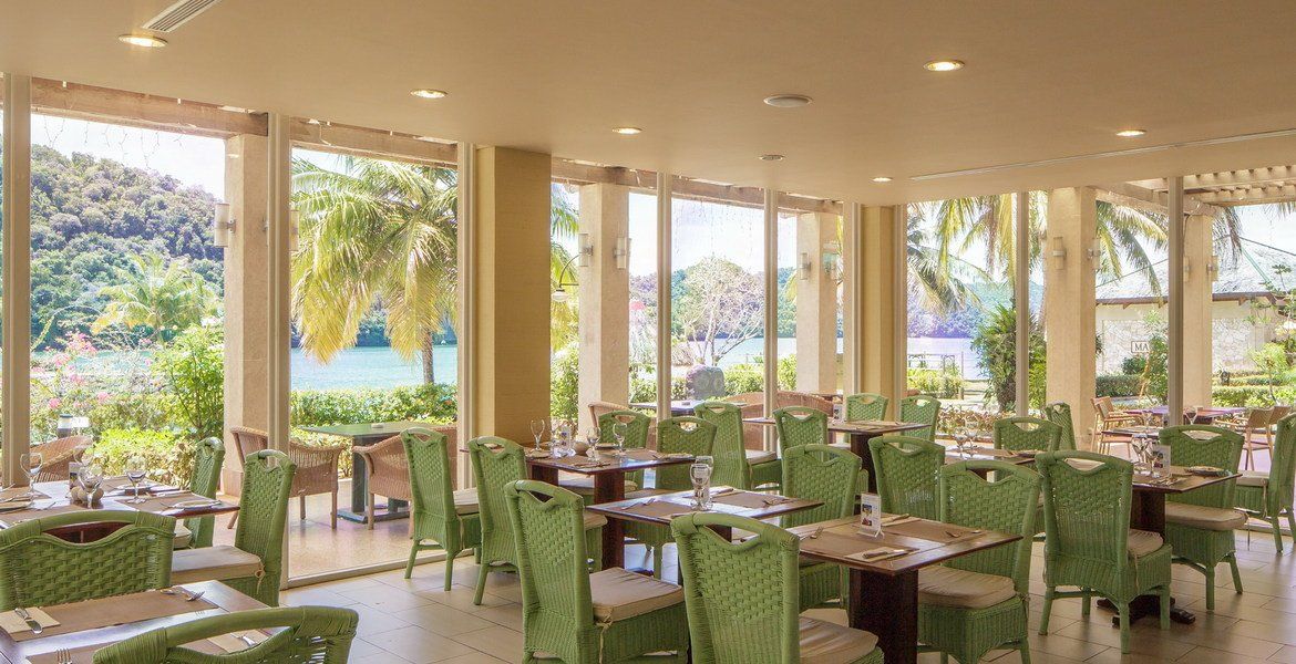 Dining room with green chairs, tables, and large windows overlooking a tropical landscape.