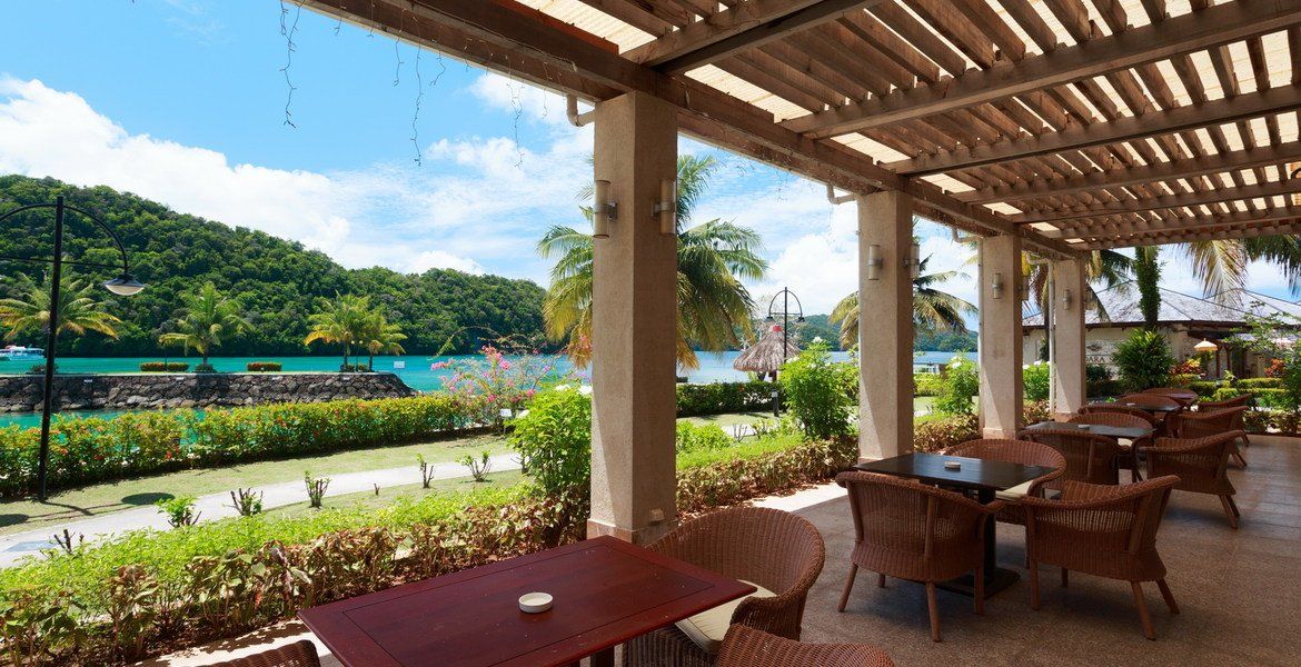 Outdoor restaurant with tables and chairs, overlooking a bay with greenery and a blue sky.