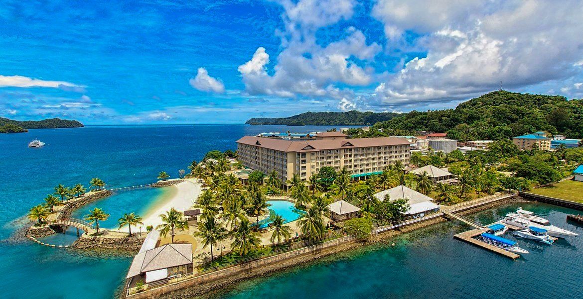 Tropical resort on blue water, sandy beach, swimming pool, boats docked, lush green hills, and blue sky.