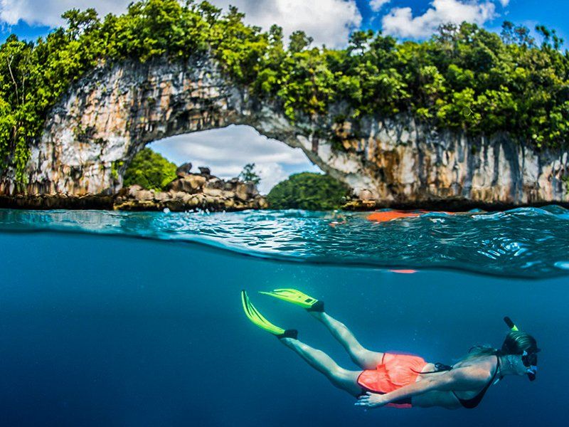 Snorkeler swims underwater near a rocky archway with lush greenery; tropical setting.