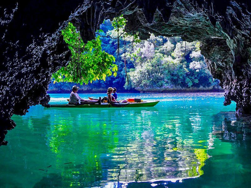 Kayakers in a green boat on turquoise water, viewed from a dark cave opening, lush island in the background.