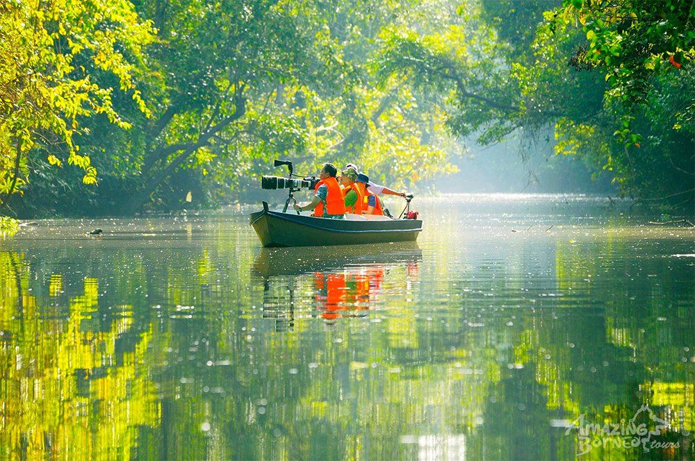 Boat with two people in orange vests on calm water, surrounded by green trees.
