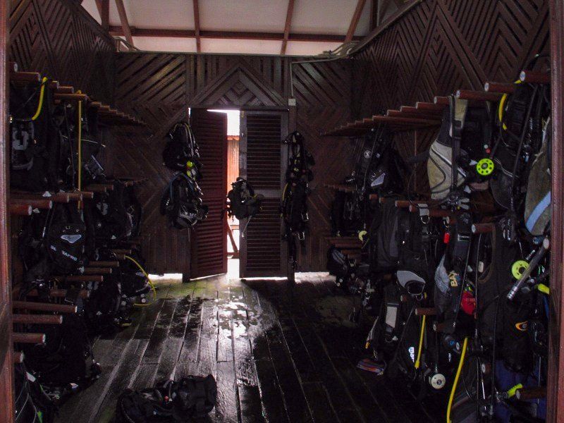 Wooden dive gear storage room; rows of black scuba equipment hanging; open doorway.