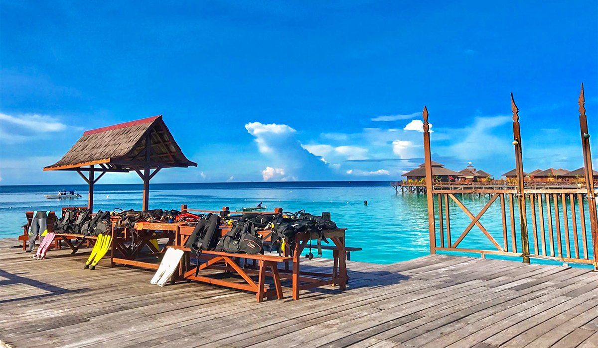 Wooden dock with scuba gear, thatched roof shelter, turquoise water, and stilt houses under a blue sky.