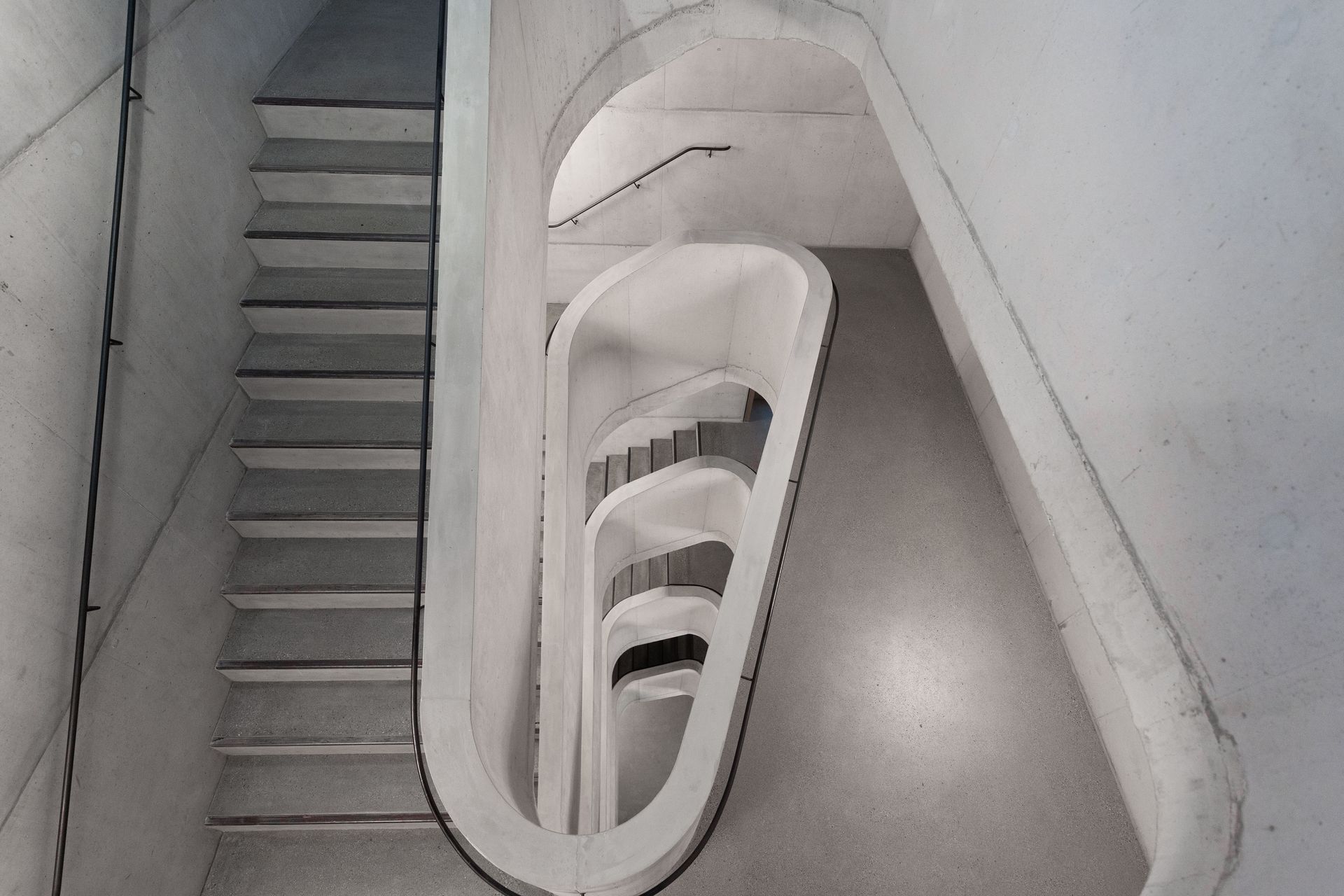 Blick in ein modernes Beton-Treppenhaus mit geschwungenen Geländern und mehreren Ebenen.