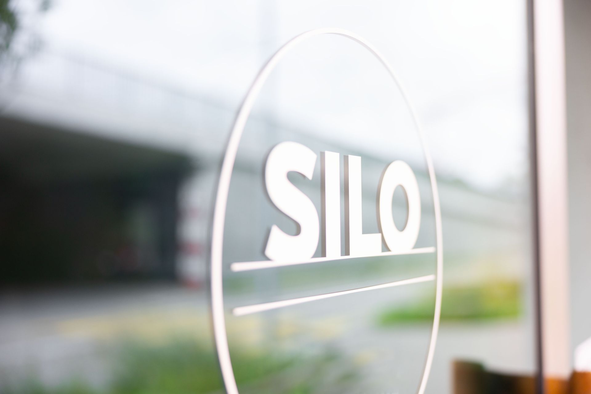 Logo „SILO“ auf einer Glastür mit unscharfem Hintergrund einer Straße und grüner Vegetation.