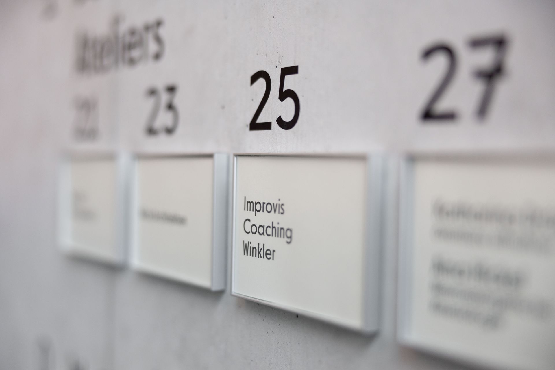 Türschild „Improvis Coaching Winkler“ neben anderen Türschildern auf einer Betonwand.