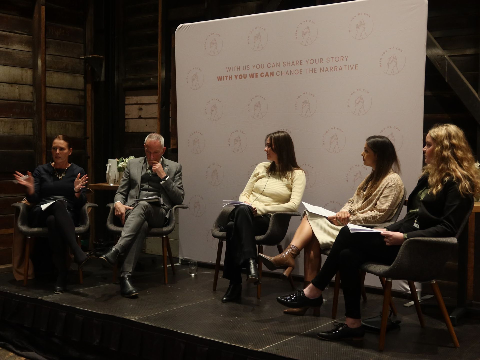 Five people sit in chairs on a stage during a panel discussion in front of a white backdrop with logo patterns.