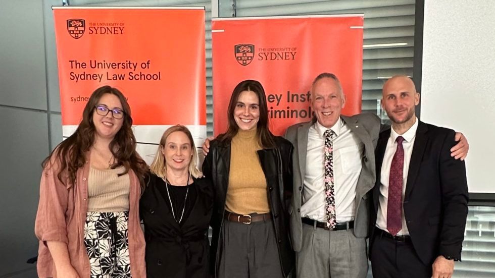 Five people stand smiling in front of University of Sydney Law School and Institute of Criminology banners.