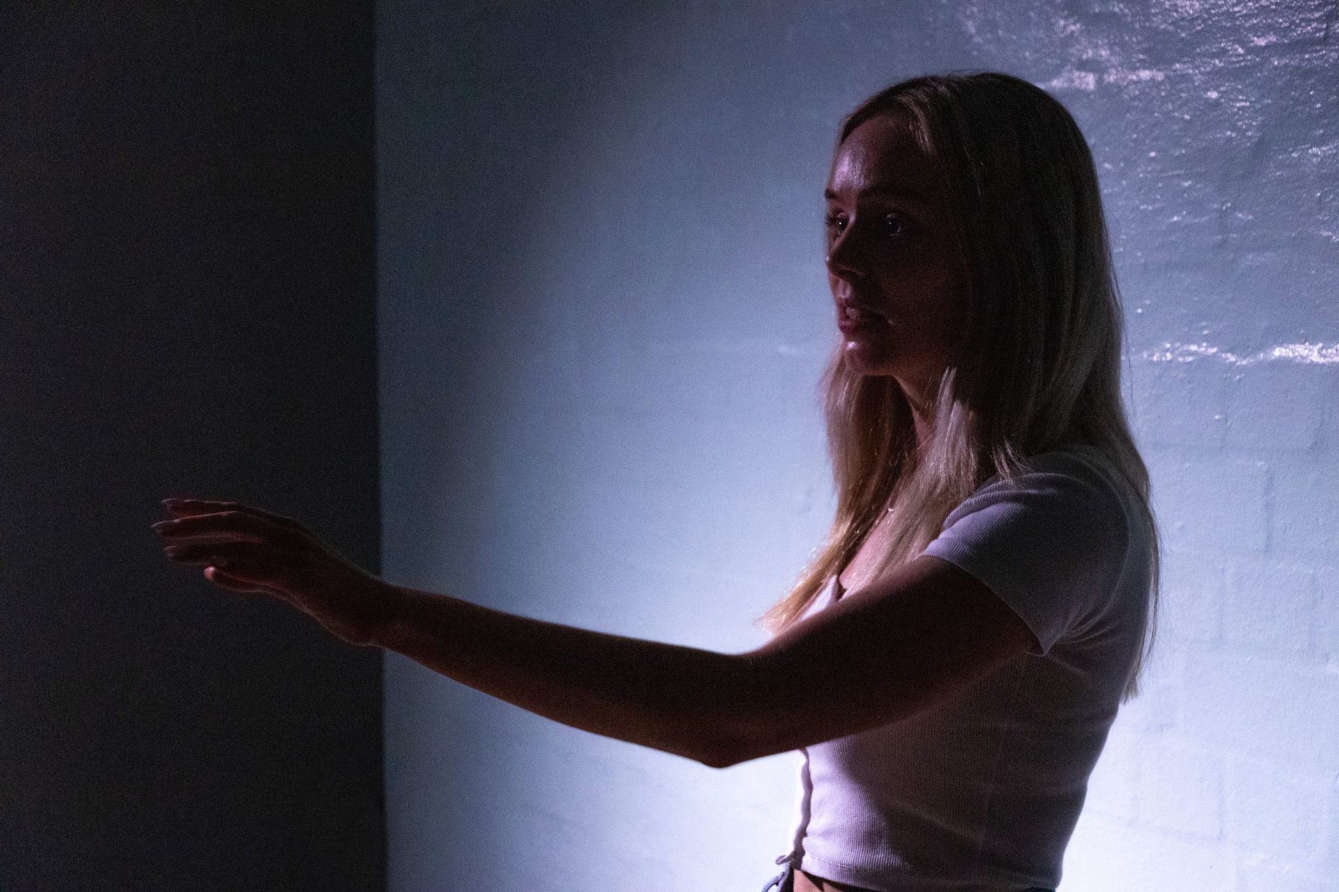 A person in a white top stands in profile, reaching out with one arm against a dimly lit blue wall.