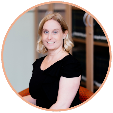 Headshot of a smiling woman with blonde hair wearing a black top, set against a blurred office background.