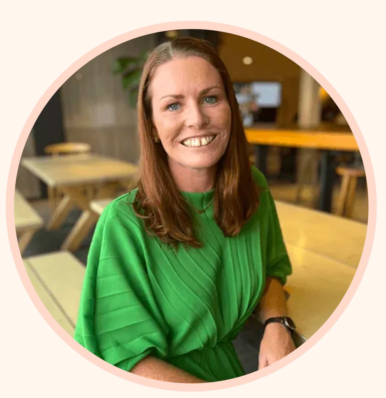 A smiling person with auburn hair wearing a bright green top, seated at a table in a blurred indoor setting.