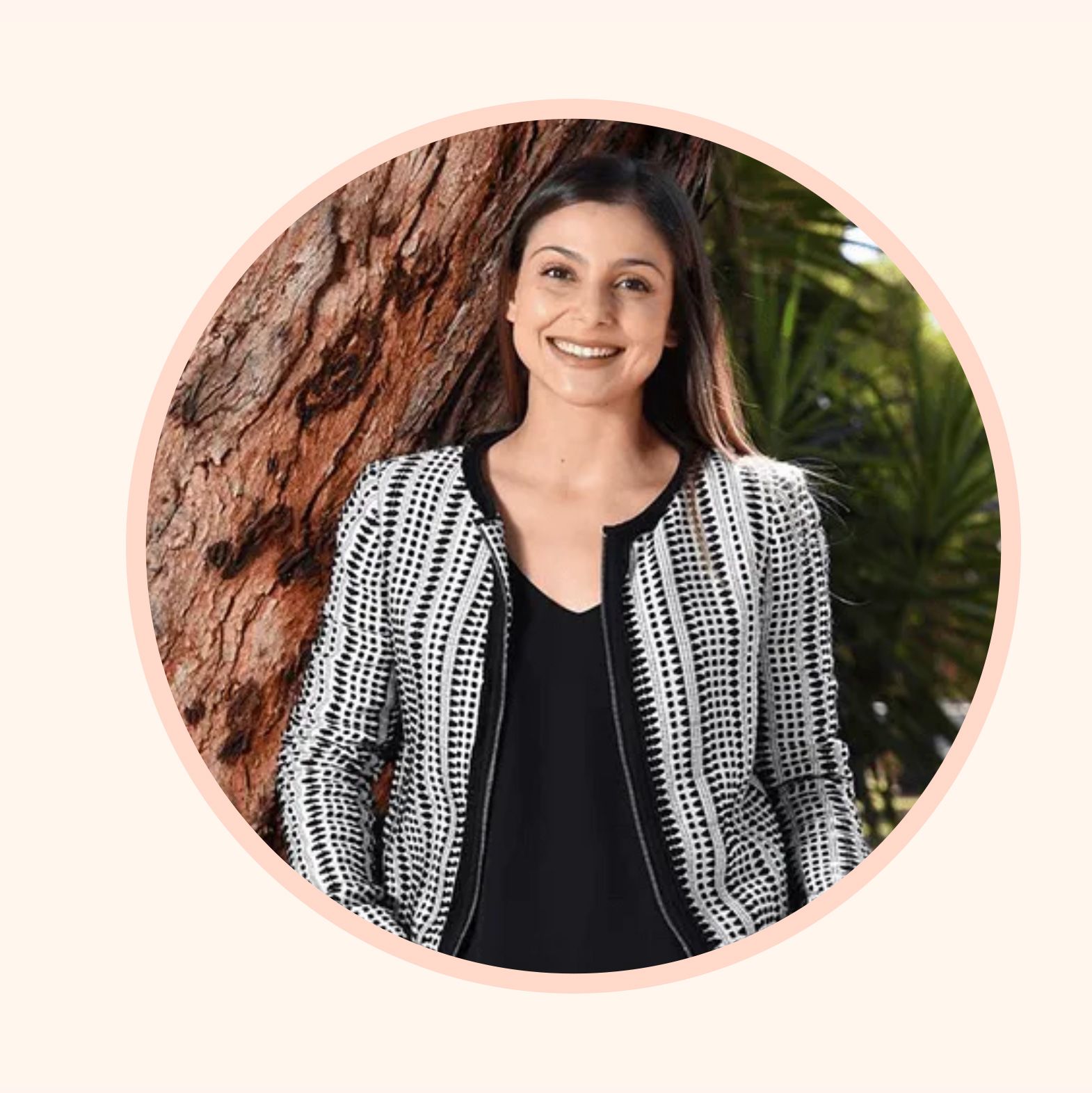 A smiling person wears a black and white textured jacket, leaning against a tree in a sunlit, leafy outdoor setting.