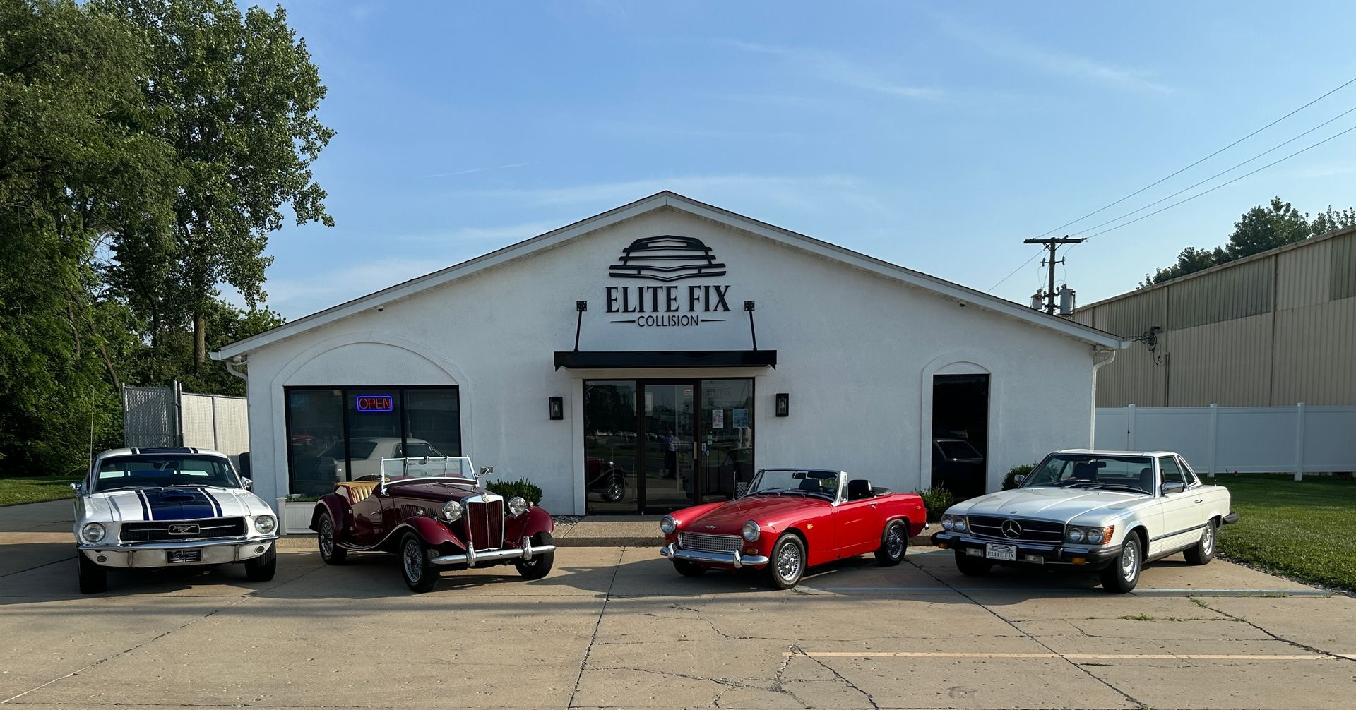 Classic cars parked in front of the Elite Fix auto shop under a blue sky.