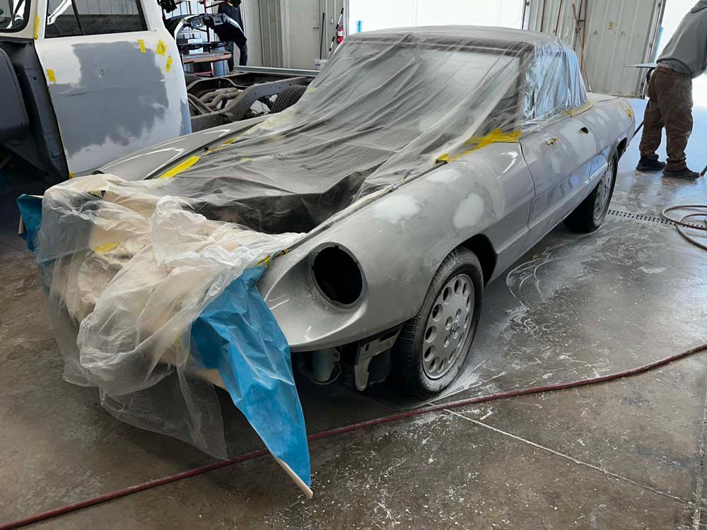 A silver car is covered in plastic while being painted in a garage.