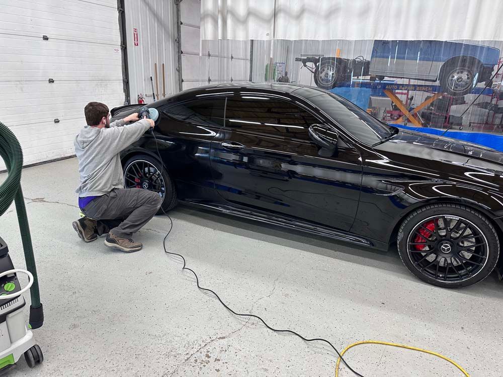 Man kneeling beside a black car in a garage, applying tint to the side window.
