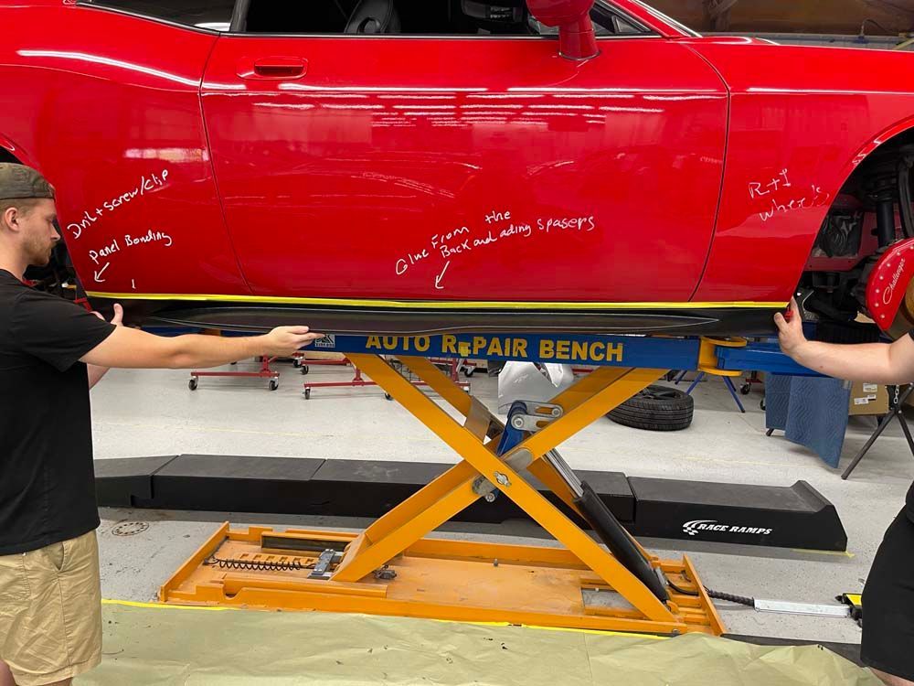 Two men are working on a red car on an auto repair bench.