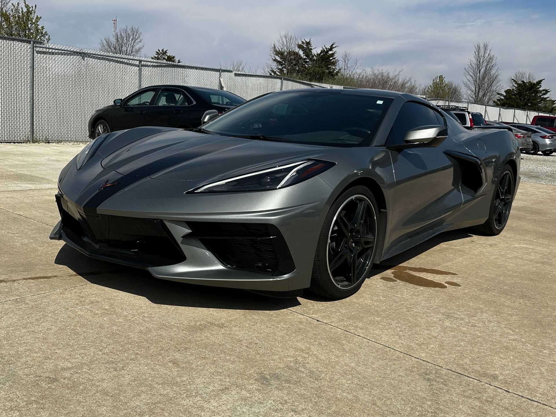 Gray Corvette sports car with black racing stripes, parked on concrete, sunny outdoor setting.