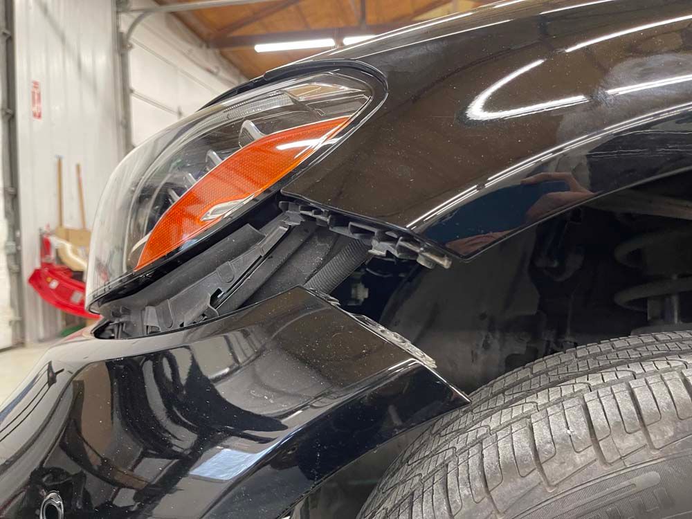 Black car's front corner with damaged bumper and headlight in a repair shop.