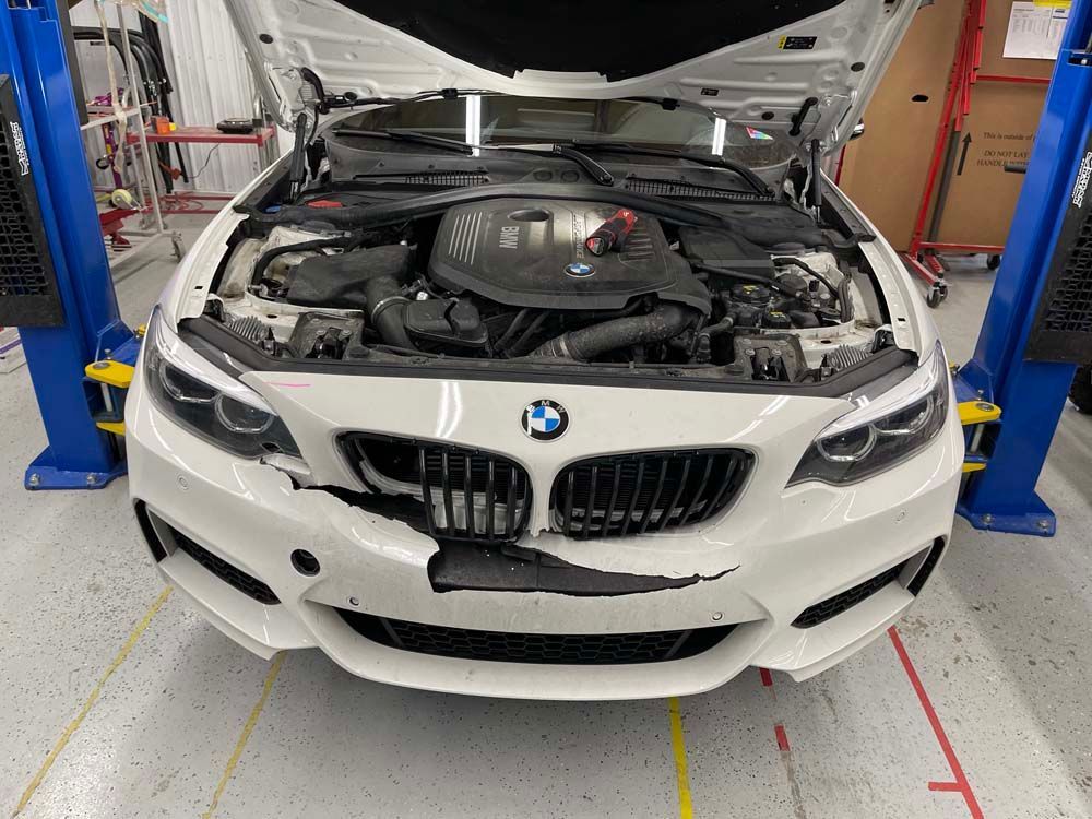 A white bmw with the hood up is on a lift in a garage.