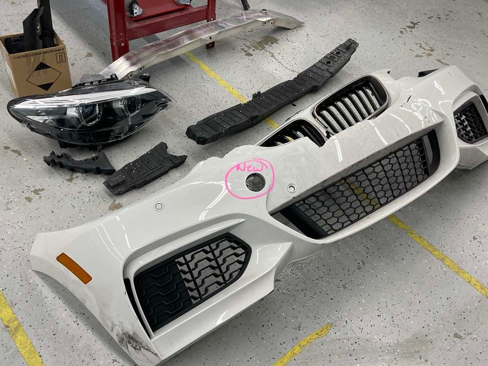 The front bumper of a white bmw is sitting on the floor in a garage.