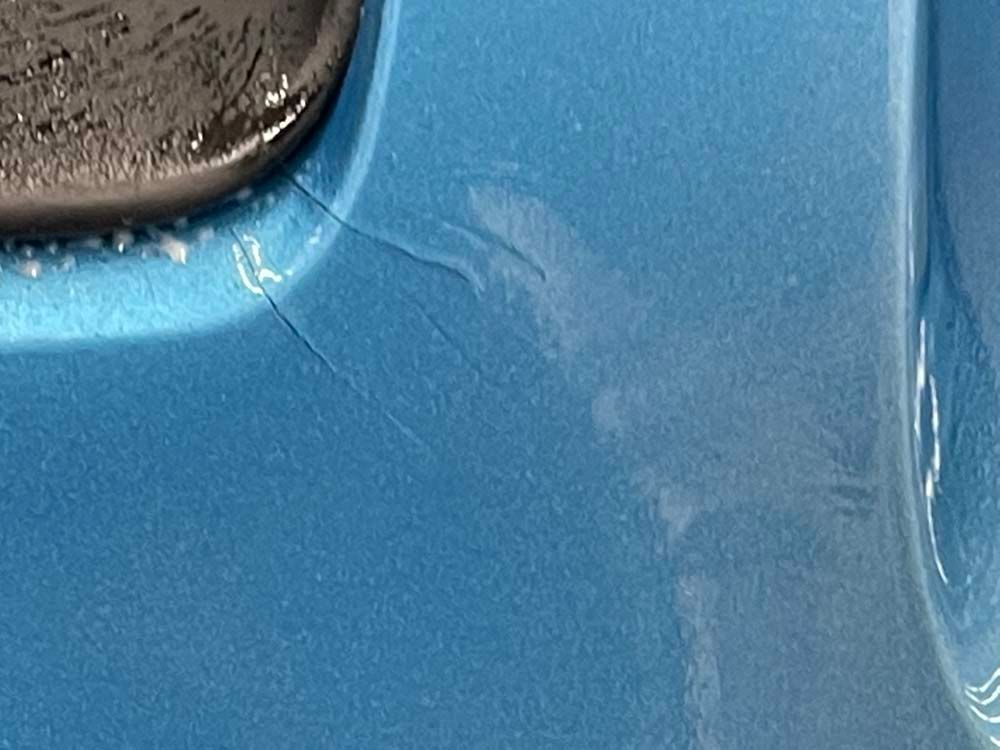 Close-up of a light blue car body with scratches and a crack near a dark, rounded object.