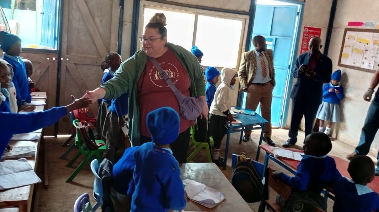 A woman in a red shirt and green jacket shakes hands with a child in a blue uniform in a school classroom. Two men in suits observe.