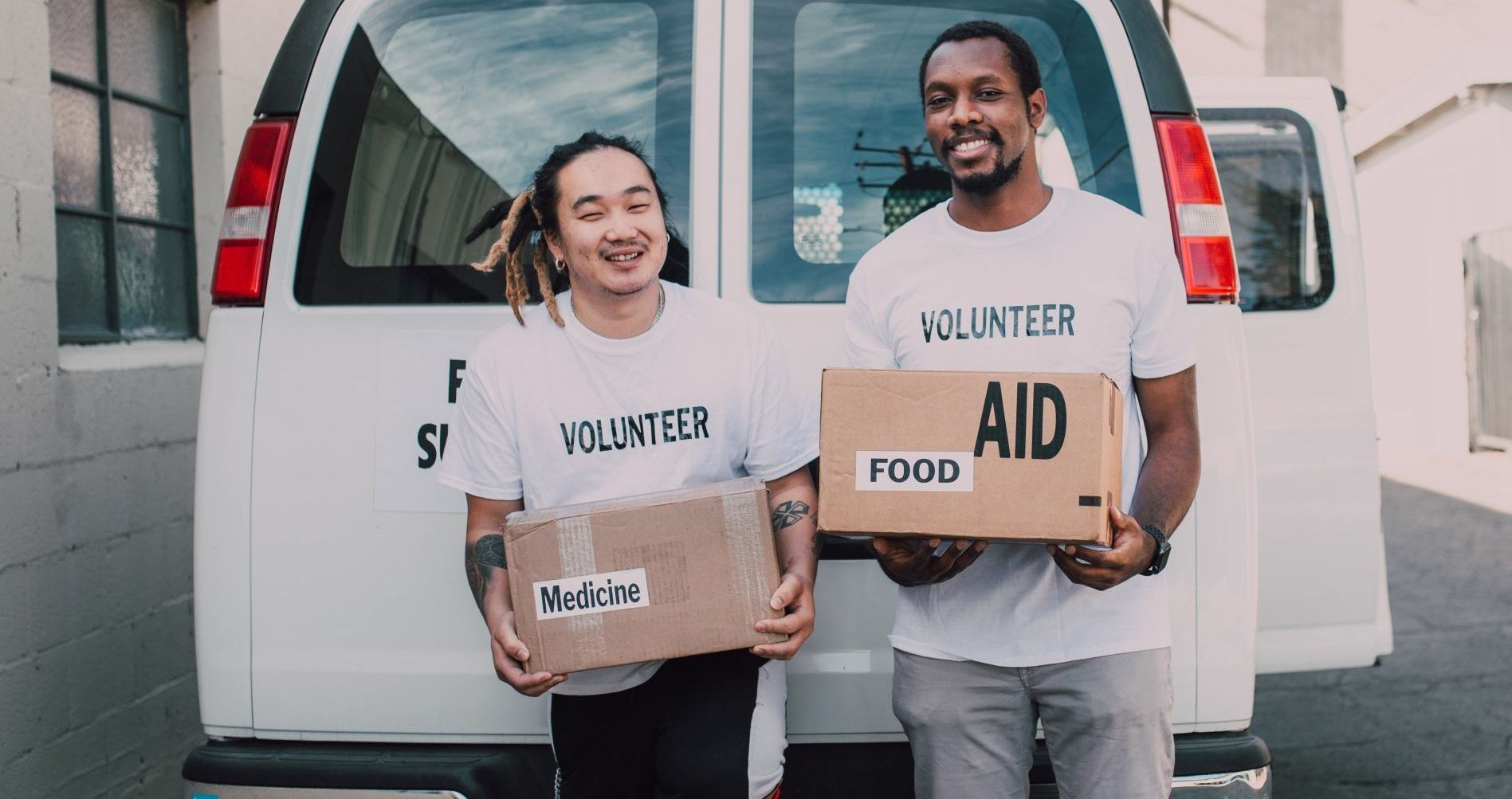 Two volunteers are holding boxes of food in front of a van.