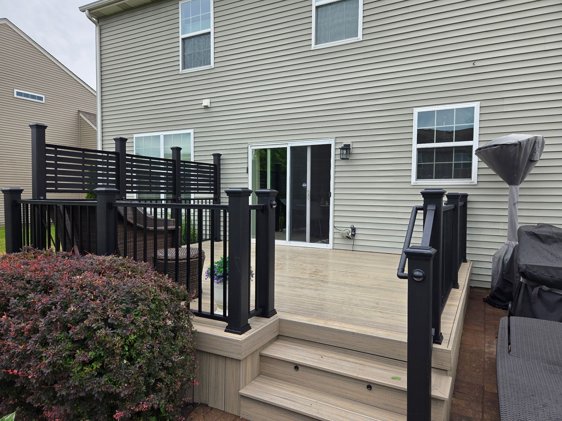 Backyard patio with steps, black railing, sliding glass door, and covered grill beside a beige house
