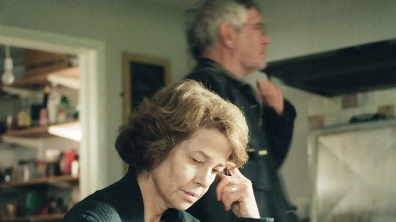 Woman with short, brown hair, looks down thoughtfully, hand to her face. An older man stands in the background, out of focus. Indoors.