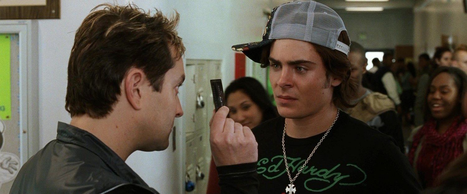 Two men in a hallway, one holding something up. The man on the right wears a backwards cap and necklace. Other people in the background.