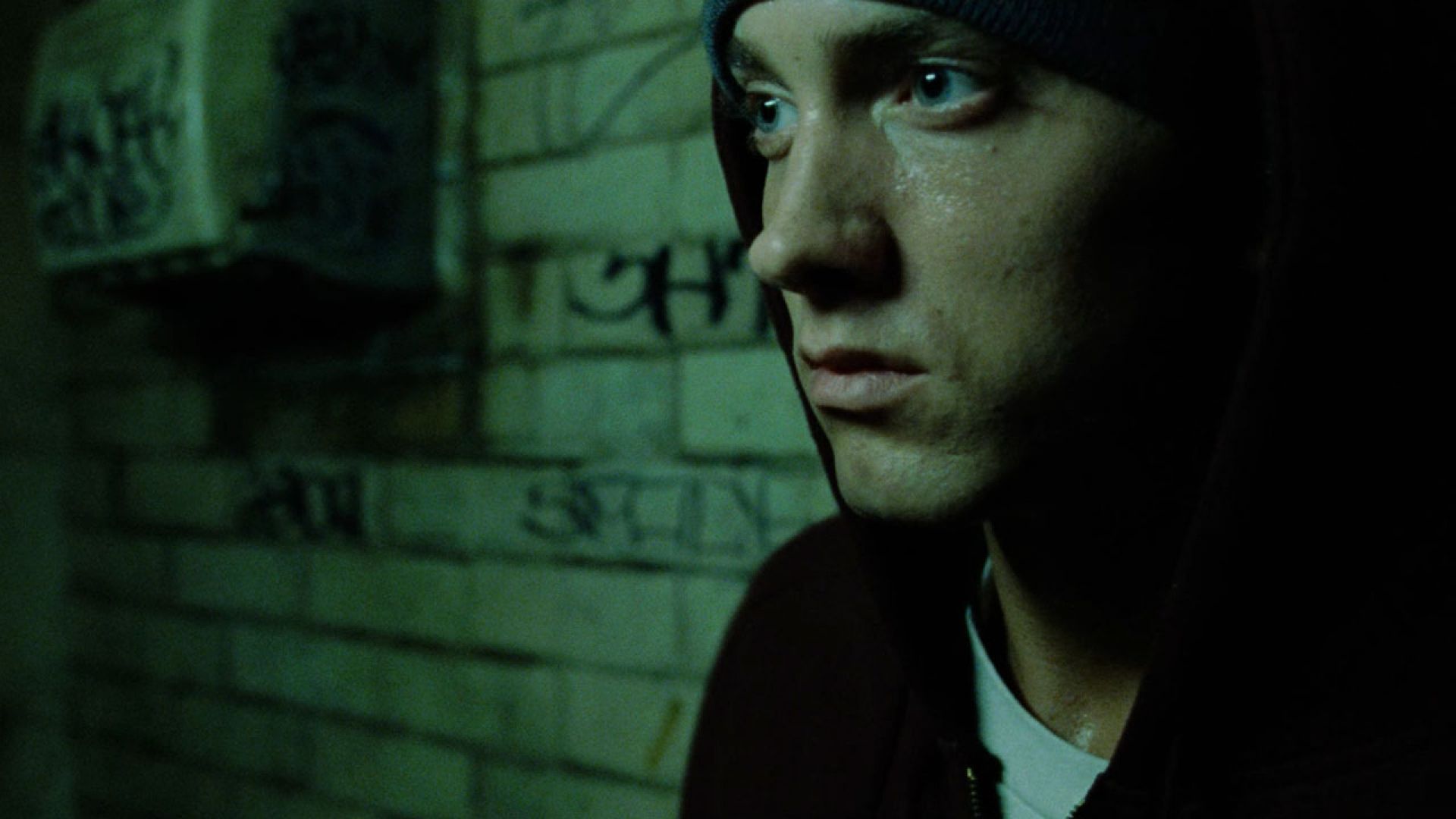 Eminem in a dark alley, wearing a hoodie, looking somber. Green-toned lighting and graffiti on the brick wall.