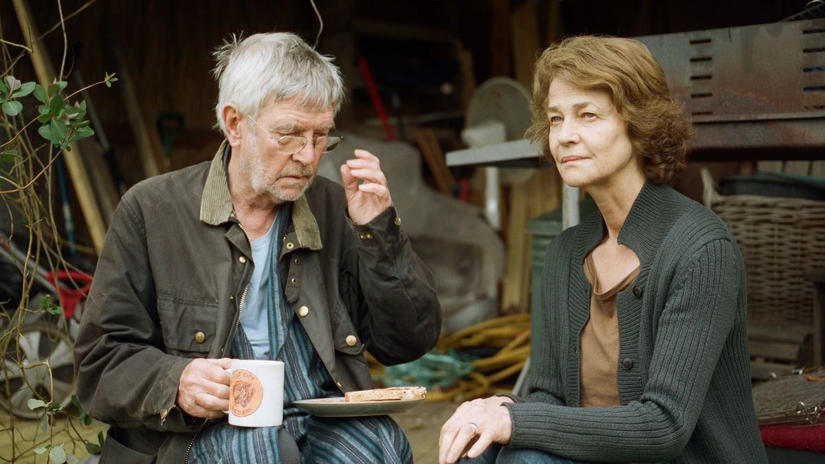 An elderly man and woman sit outdoors, eating. The man wears a jacket and holds a mug, while the woman wears a sweater.