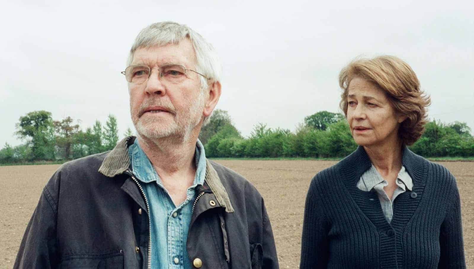 An older man and woman stand in a plowed field, looking off-camera. The man wears a dark jacket over a denim shirt, and the woman wears a blue sweater.