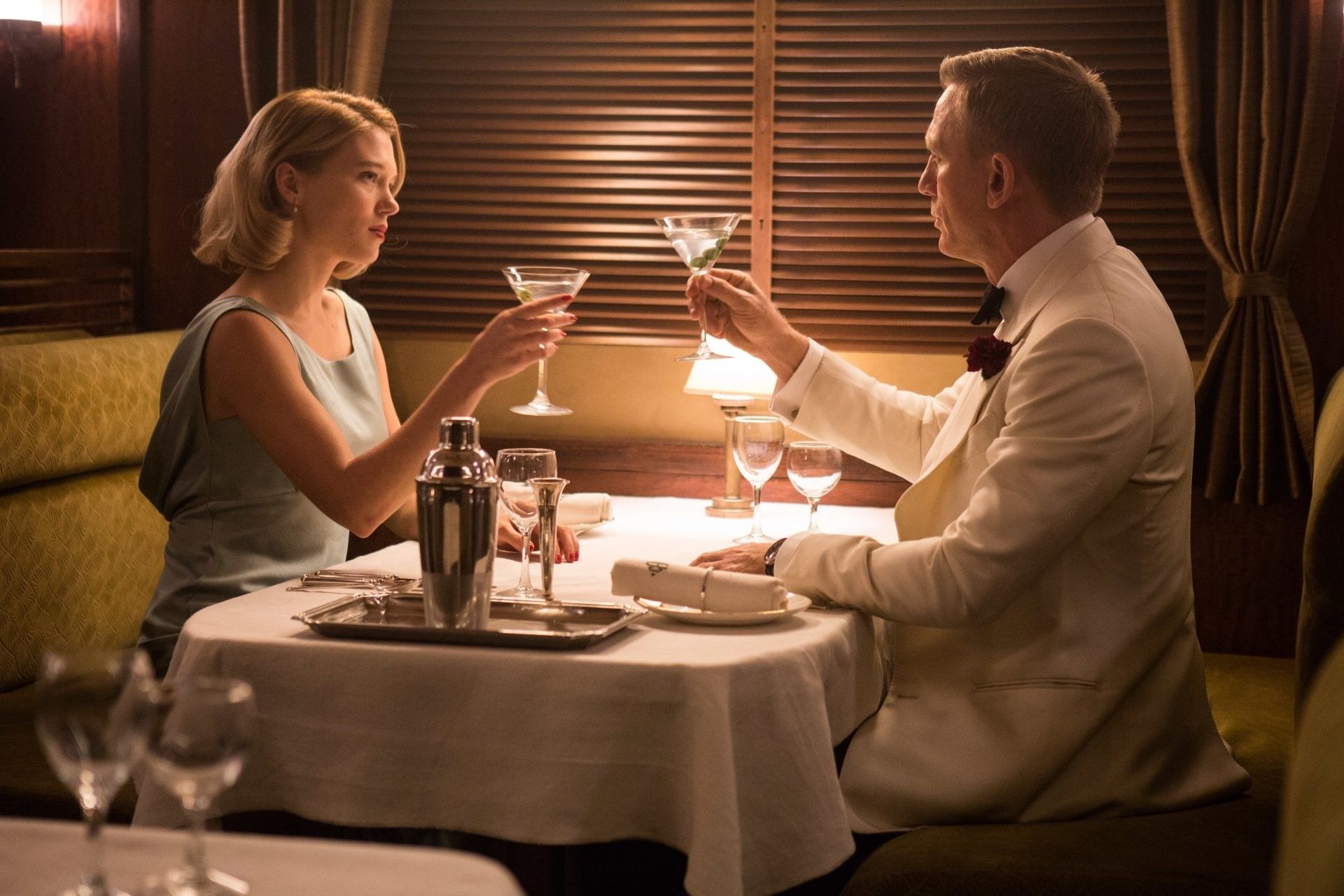 A man and woman in a luxurious dining car toast with martinis. The man wears a white suit, the woman a blue dress.