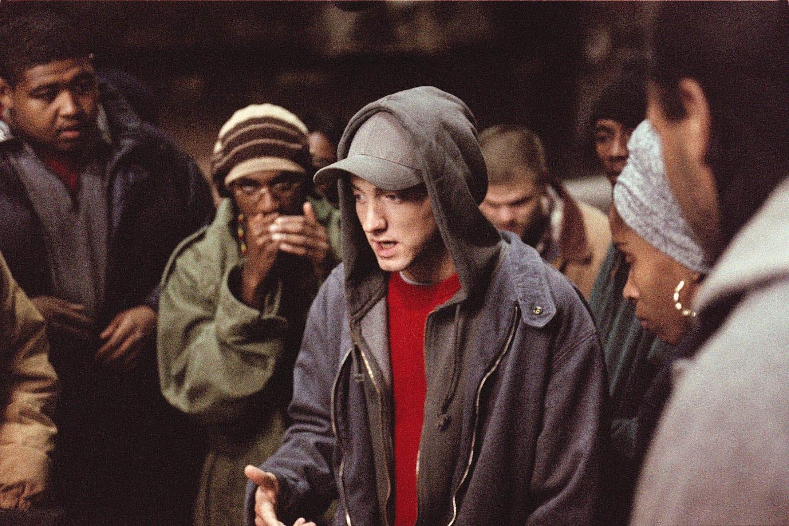Eminem, in a grey hoodie and cap, rapping to a crowd on a city street. He wears a red shirt and is gesturing with his hands.