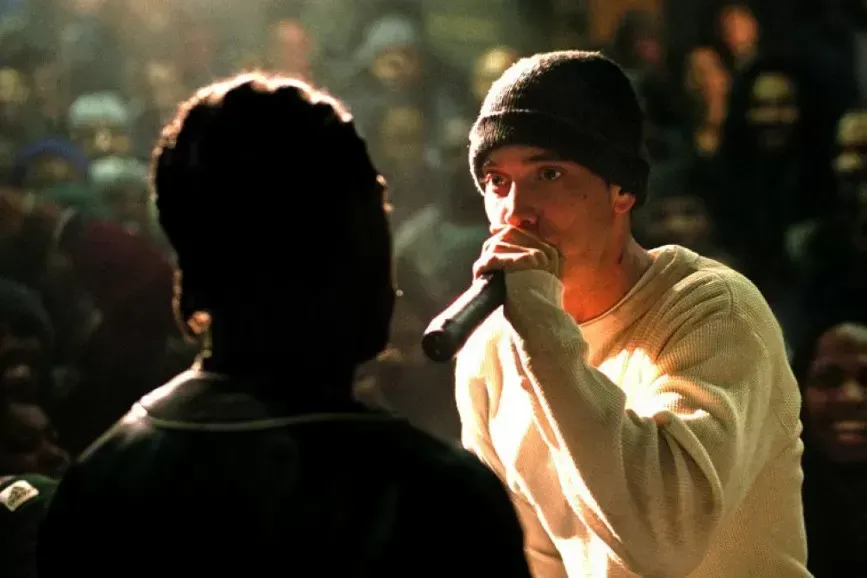 Eminem rapping into a microphone in a battle rap scene, wearing a beanie and cream-colored shirt, facing another person. A crowd is in the background.